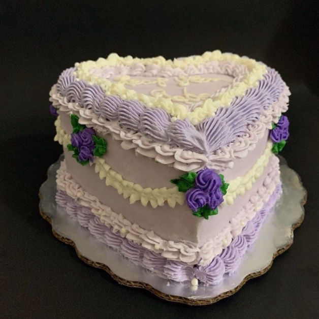 Heart-shaped cake with purple and white frosting, piped borders, small rose-like flowers on a silver board against black.