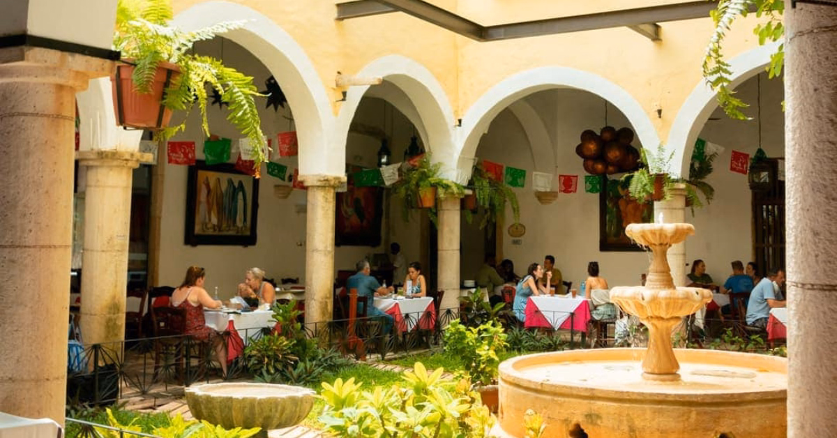 Courtyard restaurant with arched columns, hanging plants, colorful flags, and a fountain. Guests dine at festive tables.