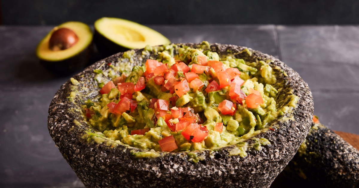 A stone molcajete with guacamole topped with chopped tomatoes on a dark surface; a halved avocado sits in the background.