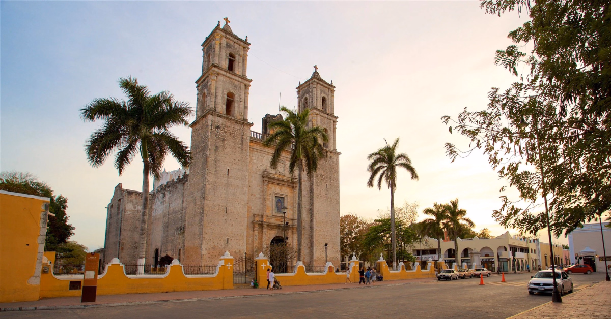 Colonial stone church with twin towers behind a yellow wall, palm trees around, people and cars nearby at sunset.