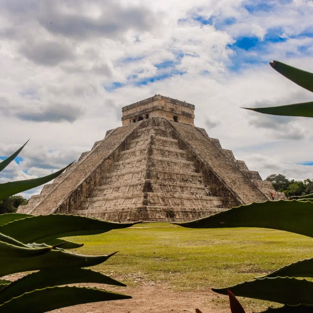 Chichen Itza’s stone pyramid rises under a partly cloudy sky, framed by green plants and surrounded by grass.