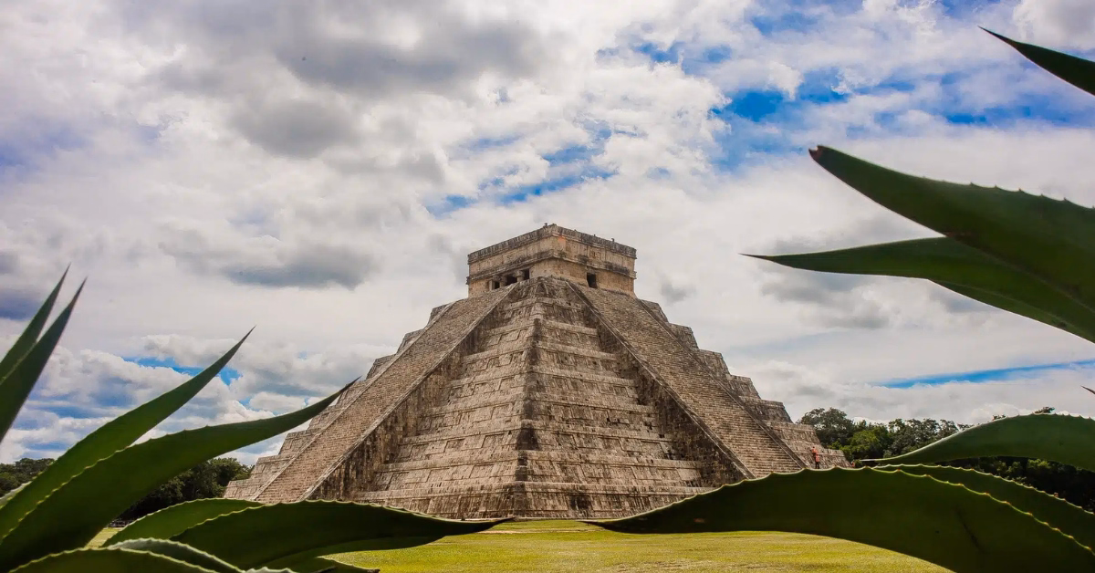 The ancient pyramid of Chichen Itza stands tall under a cloudy sky, framed by green agave plants in the foreground—a perfect view for any Chichen Itza tour.