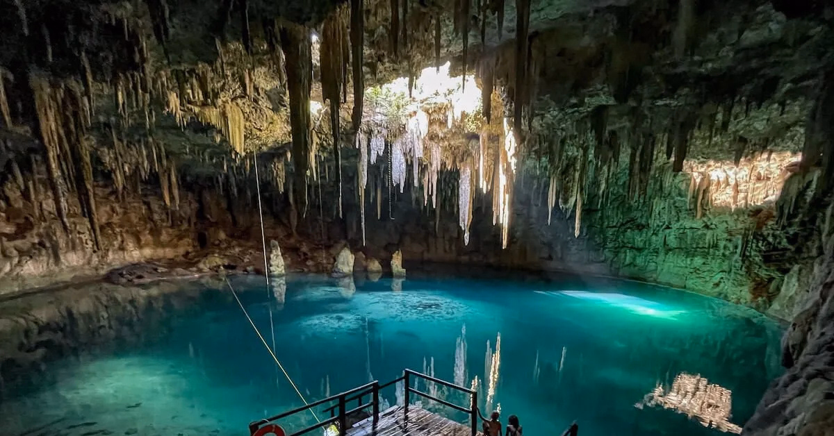 A scenic view of a clear blue underground cenote with stalactites hanging from the cave ceiling and sunlight filtering through an opening above, illuminating the water and a wooden platform—a hidden gem often visited during a Chichen Itza tour.