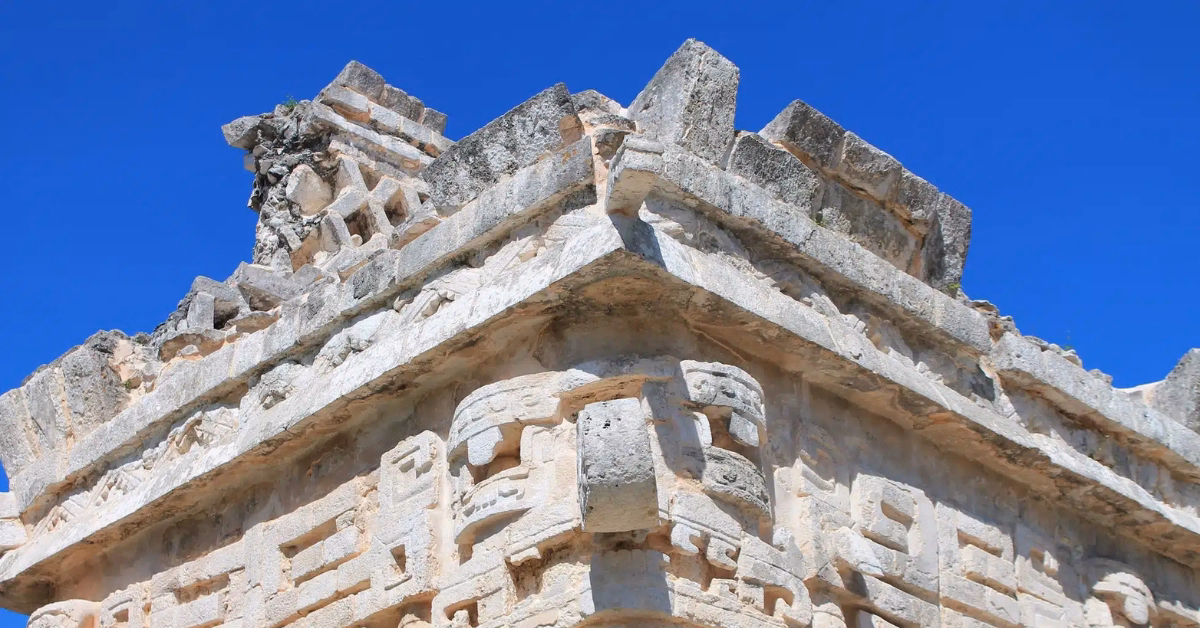 Close-up of the intricate stone carvings and geometric patterns on the upper section of an ancient Mayan temple facade against a clear blue sky, as seen during a Chichen Itza tour.