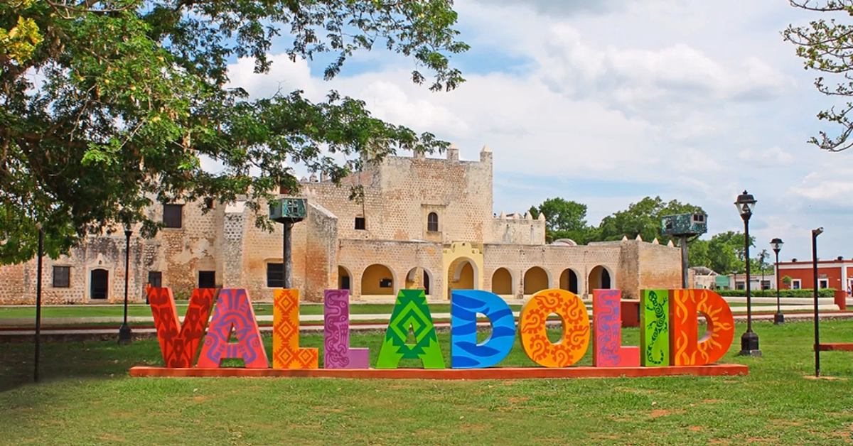 Colorful “Valladolid” sign stands before a historic stone building with arches and a tower, surrounded by green trees—an ideal stop on your Chichen Itza tour beneath the partly cloudy sky.
