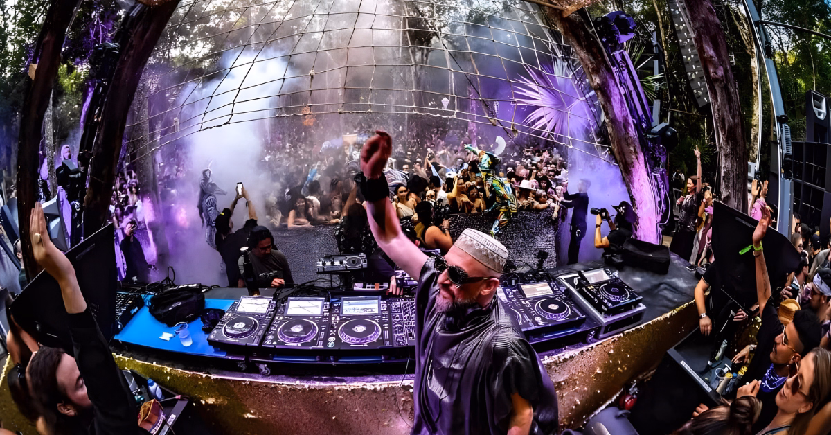 A DJ wearing sunglasses and a hat performs on an outdoor stage with multiple turntables at the Day Zero Music Festival, as a lively crowd dances and cheers under colorful lights and smoke effects in a forested area.