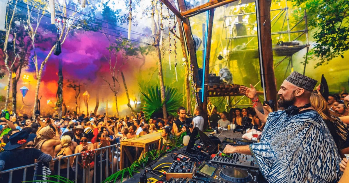 A DJ performs on an outdoor stage at the Day Zero Music Festival, surrounded by mixing equipment, while a large, vibrant crowd dances. Colorful smoke fills the air among trees, creating a festive, energetic atmosphere in the forest setting.