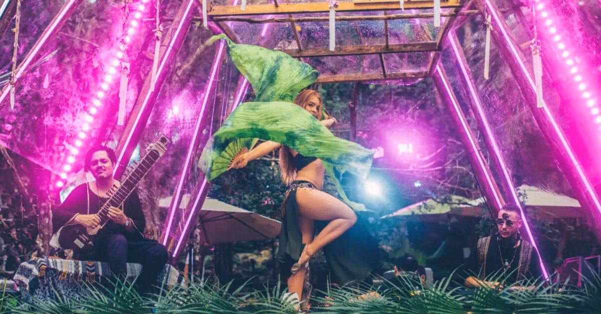A dancer twirls with flowing green fabric between two musicians, one playing a sitar, under pink lights in a lush, jungle-like setting at the Day Zero Music Festival beneath a geometric glass structure.