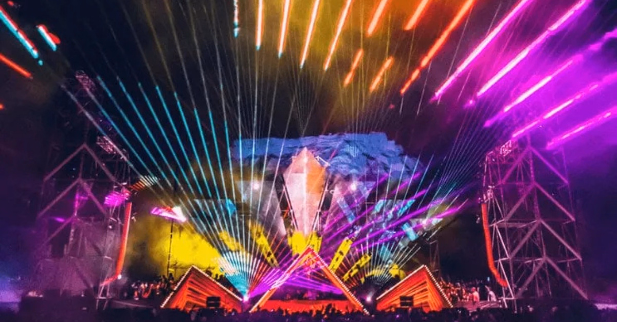 A vibrant concert scene at night with colorful laser lights and spotlights beaming across the stage and sky, geometric stage structures, and a crowd enjoying the Day Zero Music Festival.