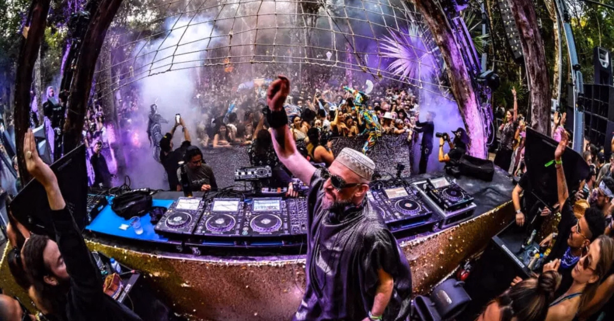 A DJ performs energetically on an outdoor stage at the Day Zero Music Festival, raising his hand as a crowd dances and cheers under colorful lights and mist, surrounded by trees.