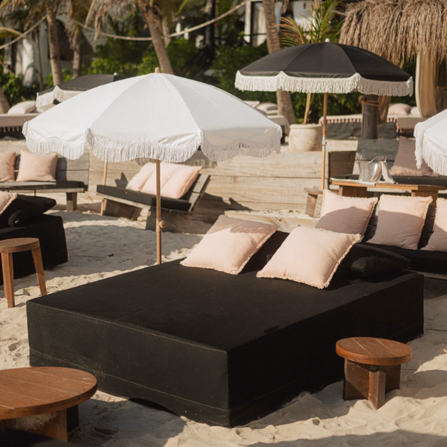 A beach lounge area at Gitano Beach Club features black sunbeds, light pink pillows, small wooden tables, and stylish white and black fringe umbrellas on sand, surrounded by palm trees.