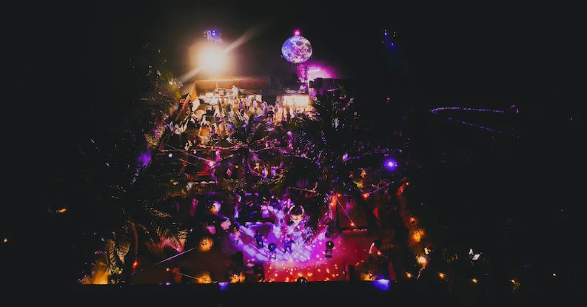 A vibrant outdoor party at night at Gitano Beach Club, illuminated by colorful lights and a disco ball, with people gathered and trees surrounding the festive area.