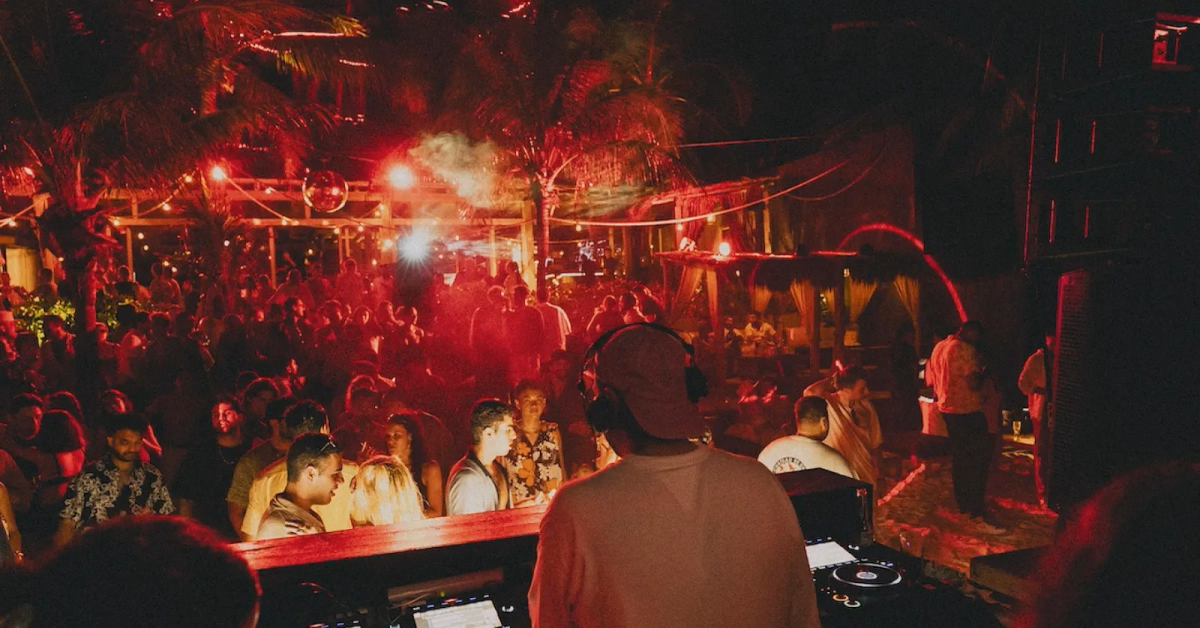 A DJ wearing headphones and a cap plays music for a large crowd at Gitano Beach Club, an outdoor nighttime party illuminated by red lights and surrounded by palm trees.