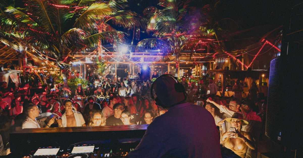 A DJ performs at an outdoor nighttime party under palm trees at Gitano Beach Club, with a lively crowd dancing and taking photos. Colorful lights illuminate the festive tropical setting.