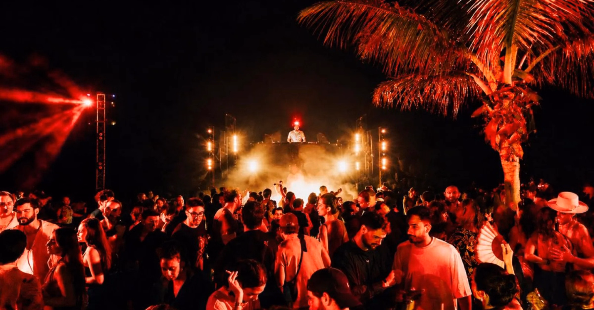 A lively crowd dances under red lights at an outdoor night party at Gitano Beach Club, with a DJ performing on a raised stage, surrounded by palm trees and atmospheric fog.
