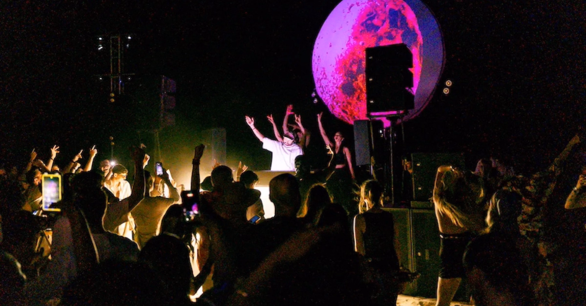 A crowd dances and raises their hands at a night concert or party at Gitano Beach Club. A performer stands on stage with arms up, a glowing orb in the background, while some capture the vibrant moment on their phones.