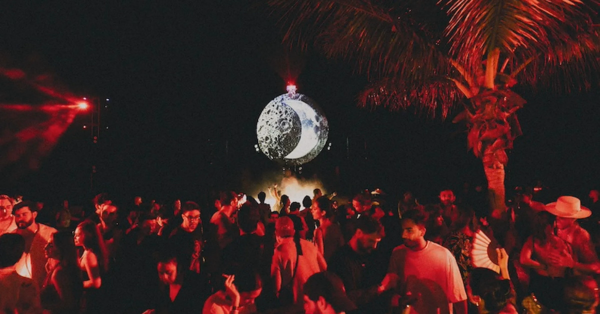 A crowd gathers at an outdoor night event under red lighting at Gitano Beach Club, with a large illuminated moon-like sphere as a central decoration and a palm tree visible to the right.