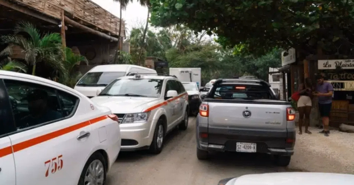 Cars and taxis in traffic on a narrow, green-lined street with small shops in Tulum; people stand nearby.
