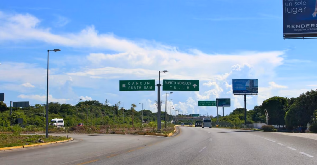 Curved highway with green signs for Cancun, Punta Sam, Puerto Morelos, and Tulum; trees, cars, and a cloudy sky behind.