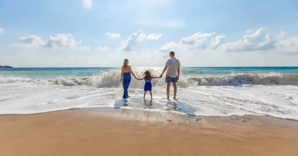 Two adults and a child stand hand in hand on a sandy Tulum beach, gazing at the ocean beneath a bright, cloud-dotted sky.