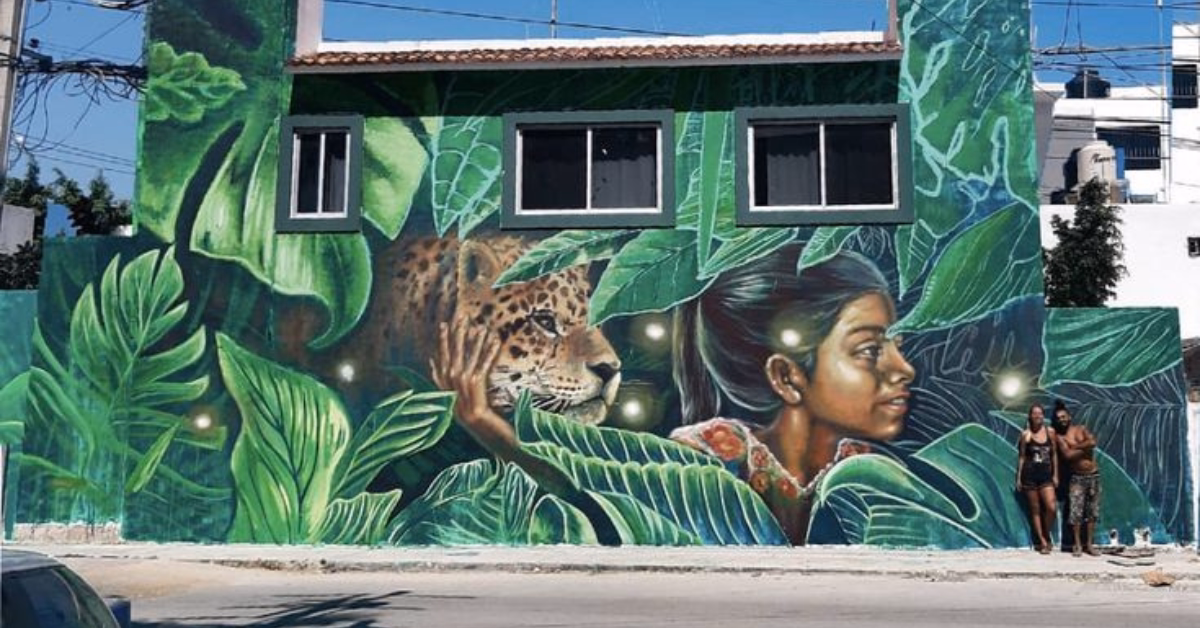 A vibrant mural on a green building, part of the Art in Tulum scene, shows a woman’s face and a jaguar amid large tropical leaves and glowing orbs. Two people stand on the sidewalk in front of the mural.