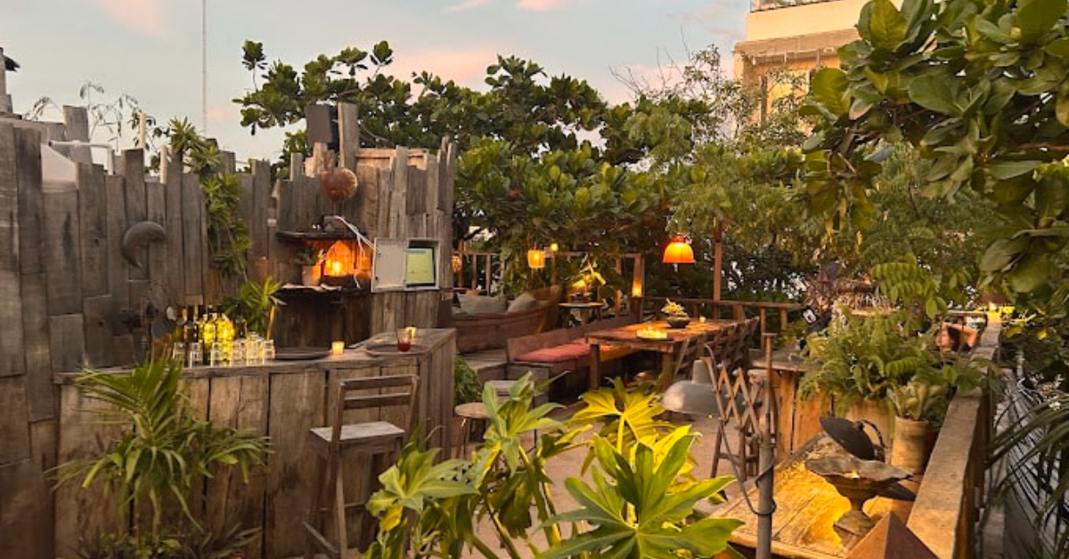 Cozy patio with rustic wood furniture, warm lights, lush plants, a small bar, and seating surrounded by trees at sunset.