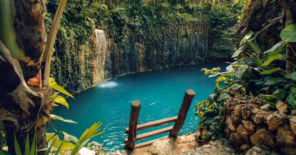A wooden stairway leads down to Cenote Atik, a natural pool surrounded by lush greenery and rocky cliffs with vines, featuring a small waterfall cascading into the turquoise water.
