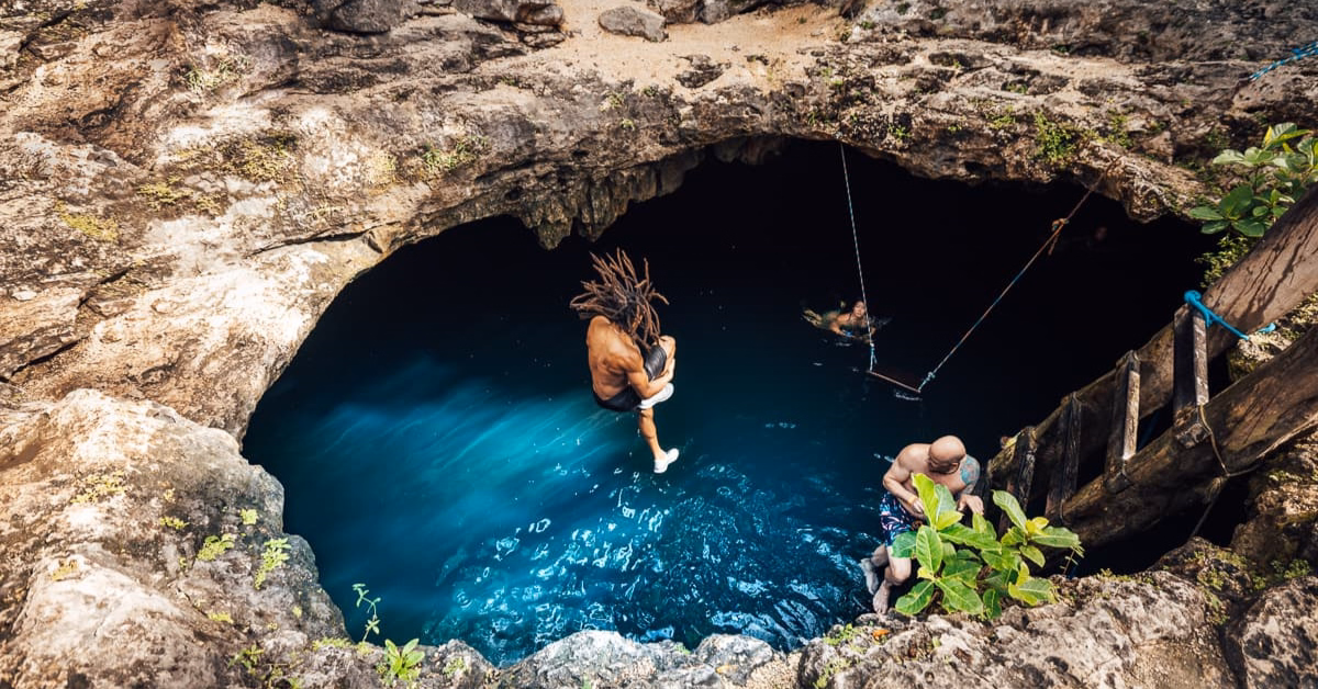 A person with dreadlocks jumps into Cenote Calavera’s deep blue water, while another stands on a wooden platform and a third swims nearby. The cenote is surrounded by rocky edges and lush greenery.