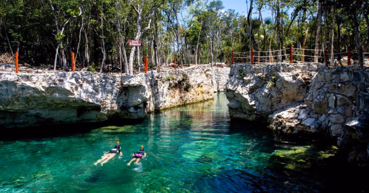 Two people snorkel in the clear, turquoise waters of Cenote Casa Tortuga, a natural pool surrounded by rocky cliffs and dense trees, with a rope fence and a sign visible in the lush outdoor setting.