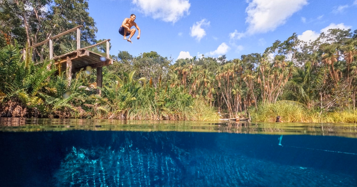 Someone leaps from a wooden platform into clear blue water at Cenote Cristal, surrounded by lush trees and a bright sky.