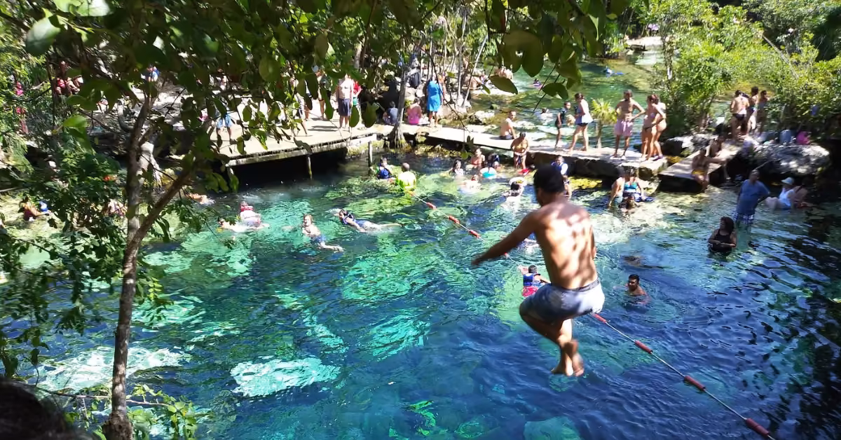 A man leaps into turquoise cenote waters as others swim or watch from a wooden boardwalk amid lush greenery.