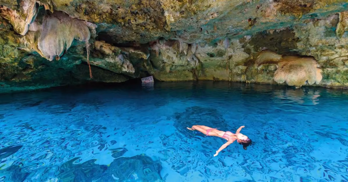 A person floats on their back in clear blue water at Cenote Dos Ojos, with rocky cave walls and a partially open ceiling above.