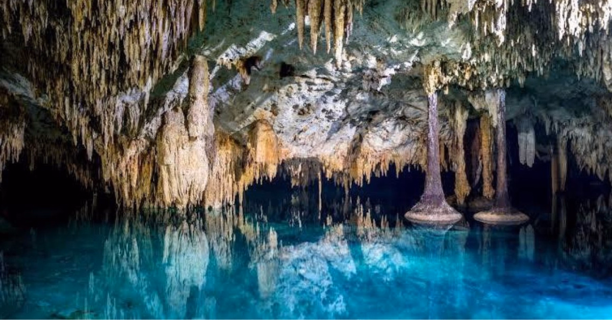 A cave with hanging stalactites, rising stalagmites, and clear blue water below reflecting the rocky formations.