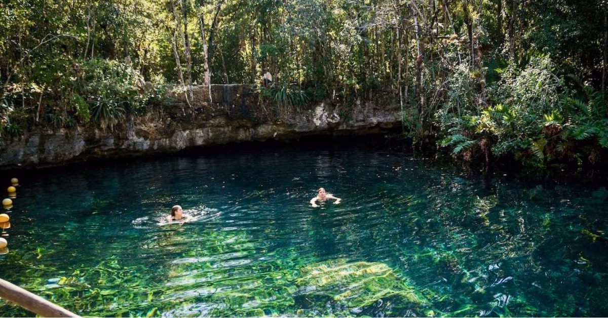 Two people swim in the clear waters of Cenote Tankah, surrounded by lush green trees and rock formations, with sunlight reflecting off the surface. Yellow buoys mark one side of the serene swimming area.