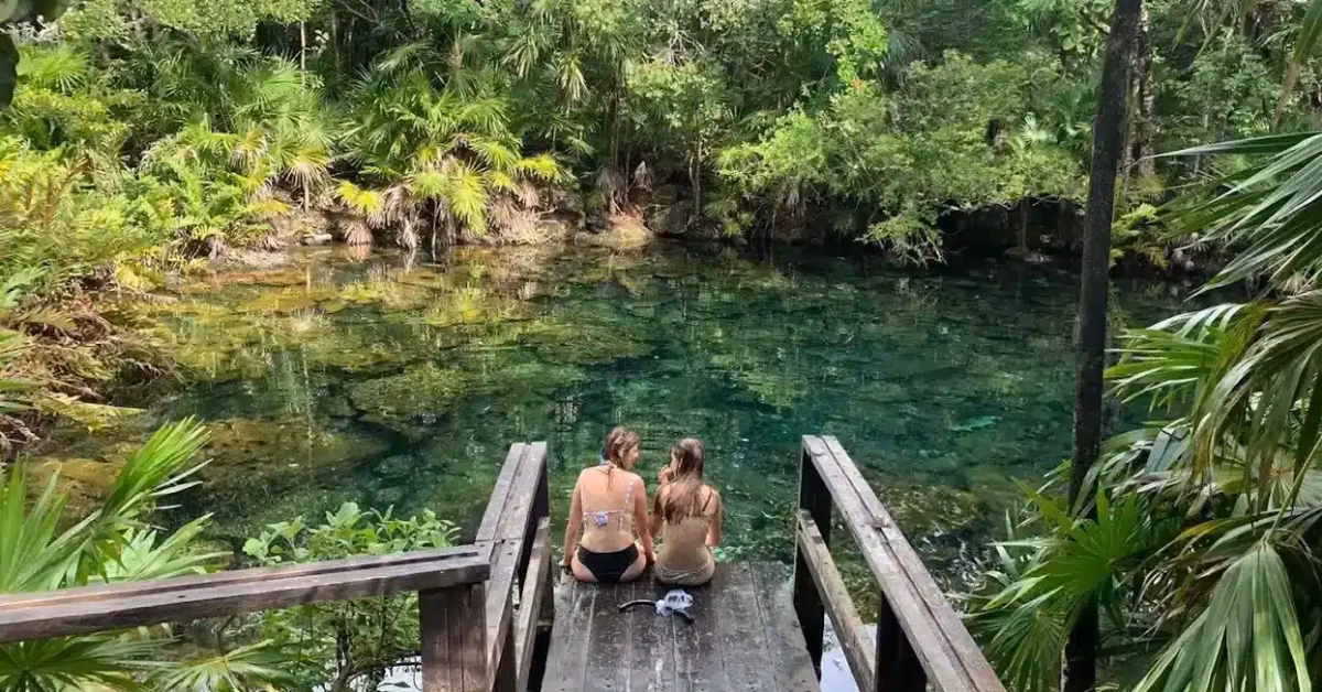 Two people in swimsuits sit on wooden steps overlooking the clear, natural waters of Cenote Tankah, surrounded by lush green tropical plants and trees.