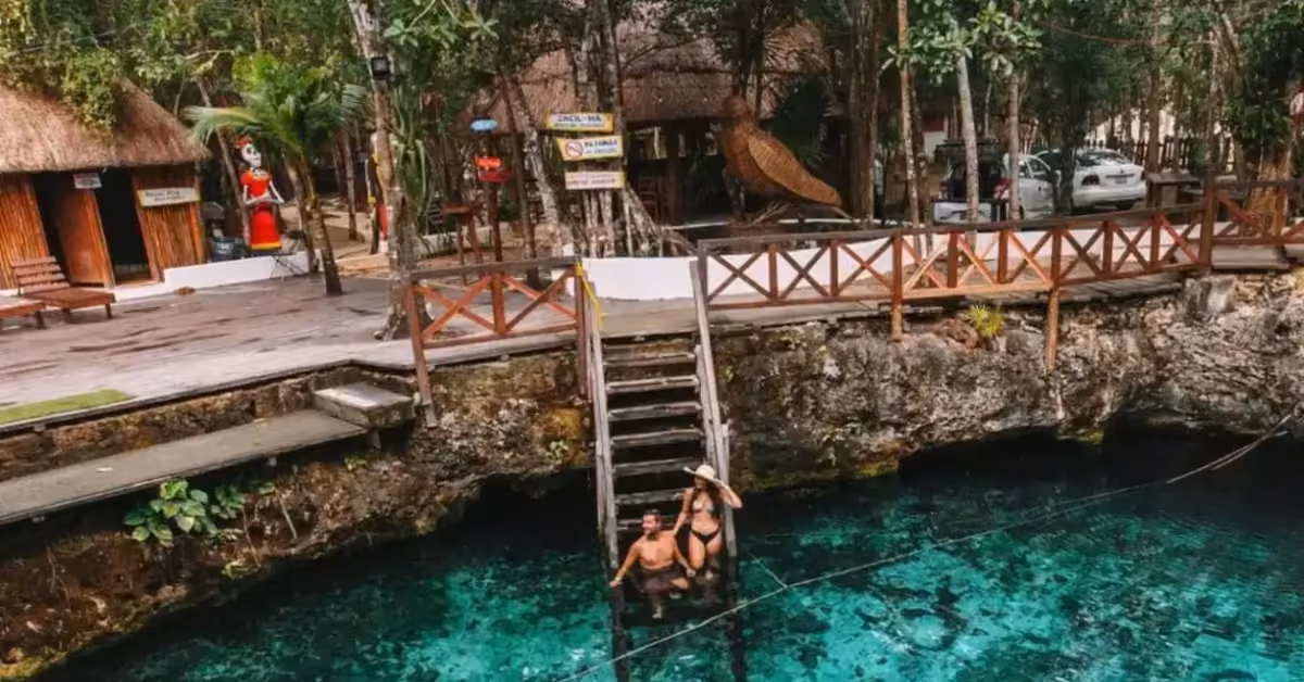 Two people sit on wooden steps above clear blue water at Cenote Zacil-Ha, surrounded by lush trees and rustic thatched buildings.
