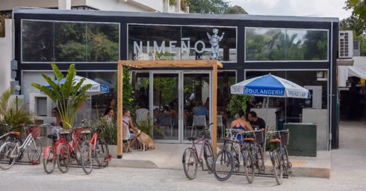 Chic black bakery with glass windows, outdoor tables under blue umbrellas, bikes parked outside, green plants, and a dog at the door.
