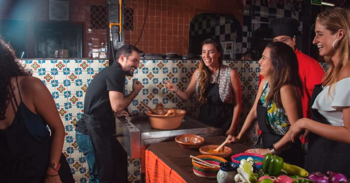 Four people in aprons laugh and cook together in a bright tiled kitchen, fresh vegetables and bowls on the table before them.