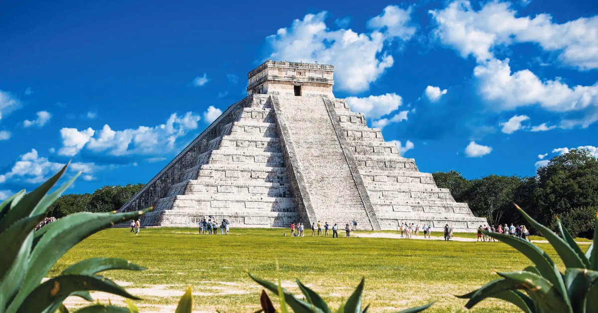 Stone pyramid of Chichen Itza under a blue sky with scattered clouds, surrounded by green grass and plants, with small groups of people near the base for scale.