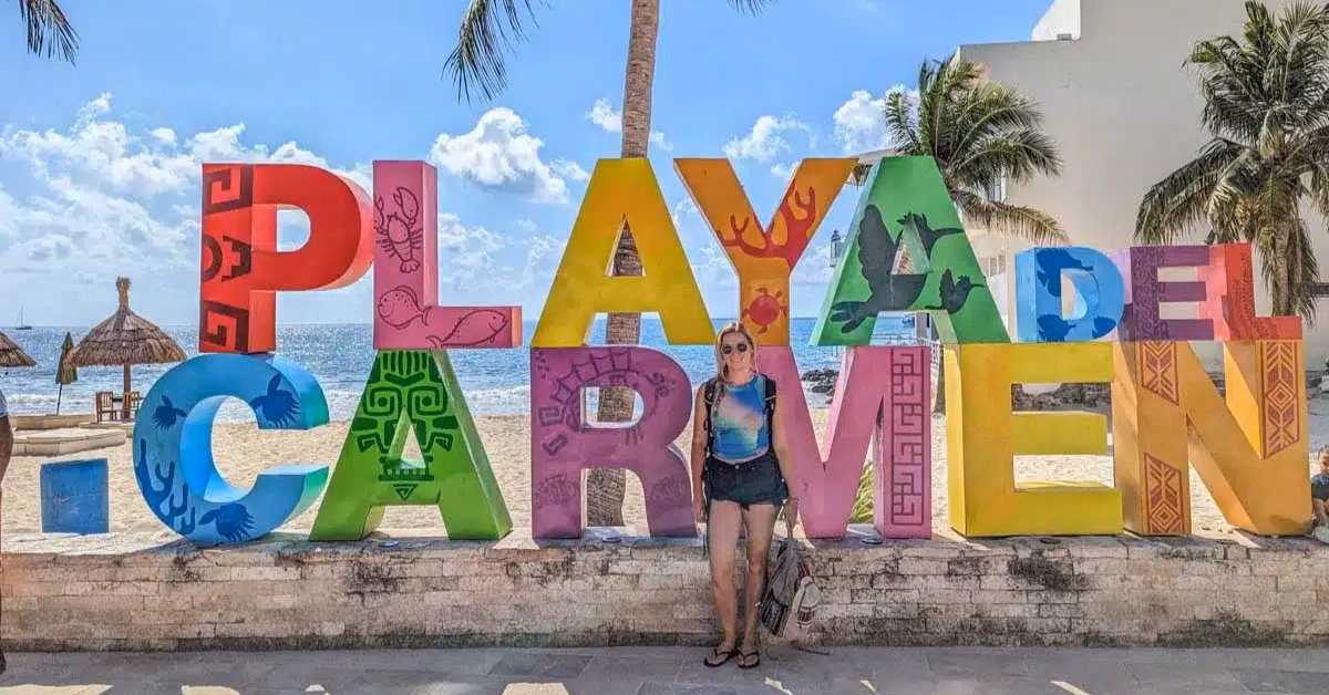 A woman stands smiling in front of a colorful Playa del Carmen sign with the beach and palm trees behind her—a perfect stop for those enjoying day trips from Tulum.