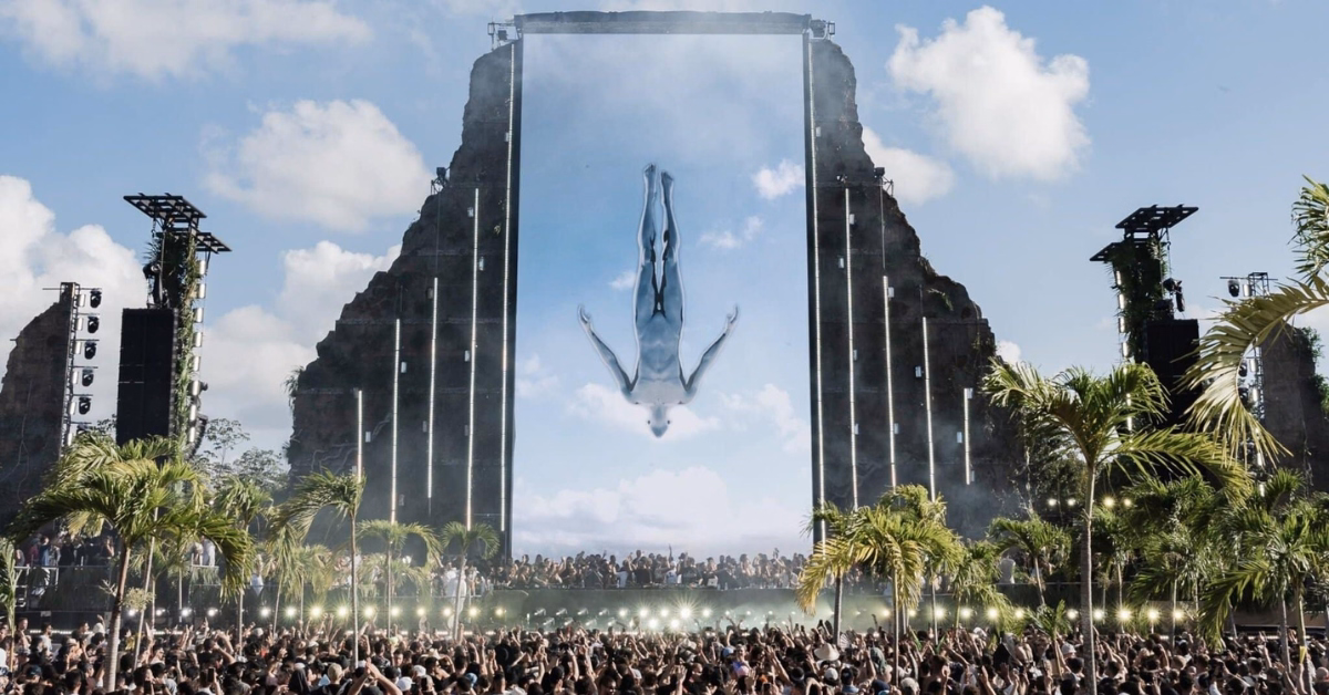 A large crowd attends an outdoor concert with palm trees and mountainous scenery, reminiscent of festivals in Tulum, while a massive vertical screen displays a digital image of a person floating upside down against a bright sky.