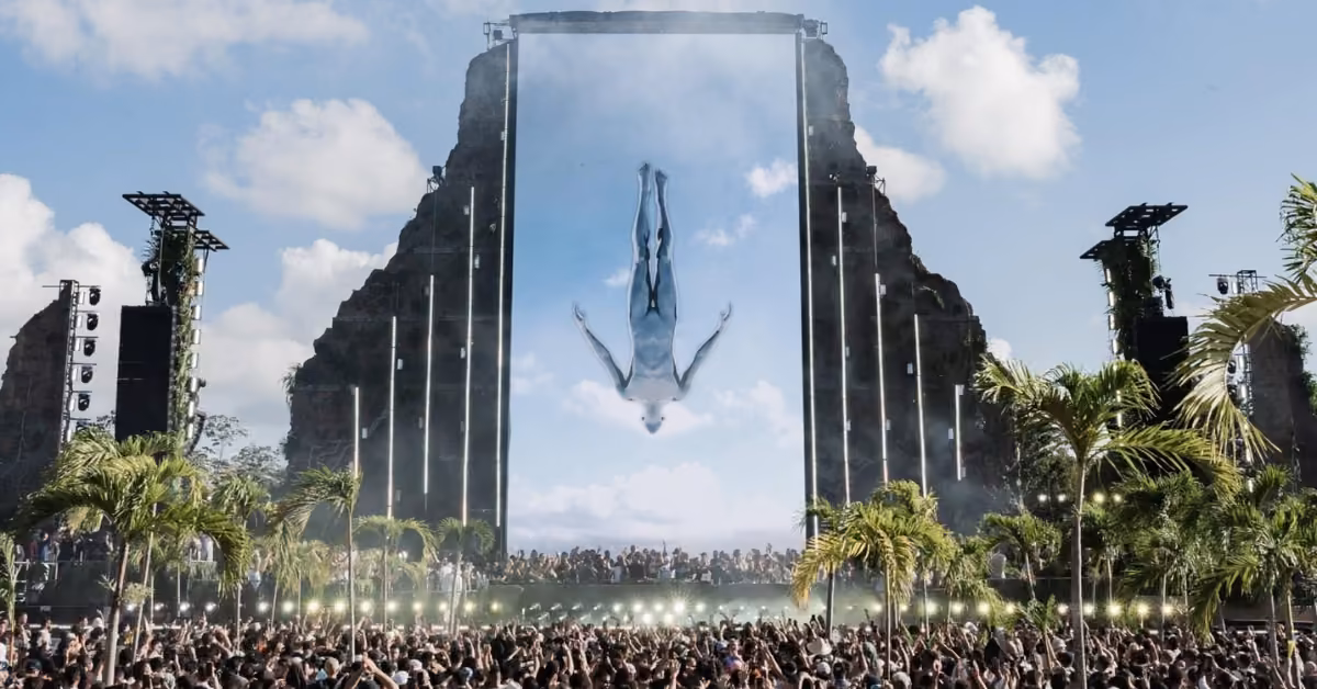Festival crowd enjoys music outdoors by a rocky stage, with diving image onscreen, palm trees and lights glowing in front.