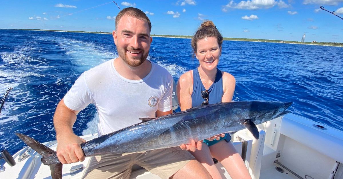 A smiling man and woman sit on a boat holding a large fish, enjoying Fishing in Tulum with deep blue ocean and clear sky in the background. The man wears a white shirt, the woman a blue tank top, and the shoreline is visible in the distance.