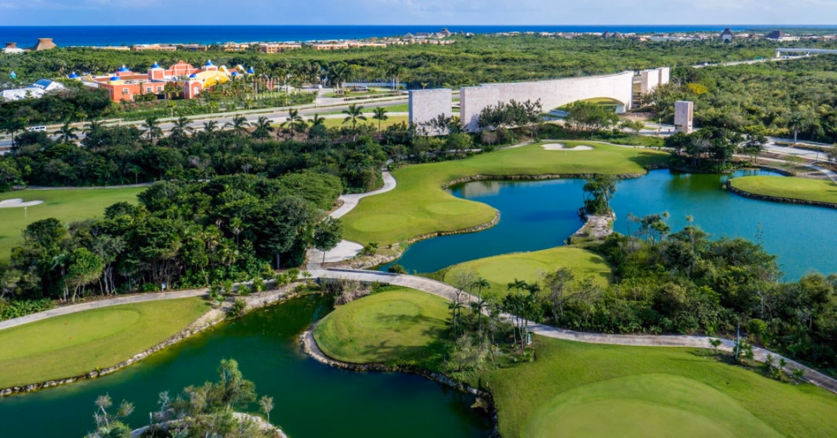 Aerial view of a lush golf course with ponds, winding paths, green landscape, and colorful buildings under a bright blue sky.