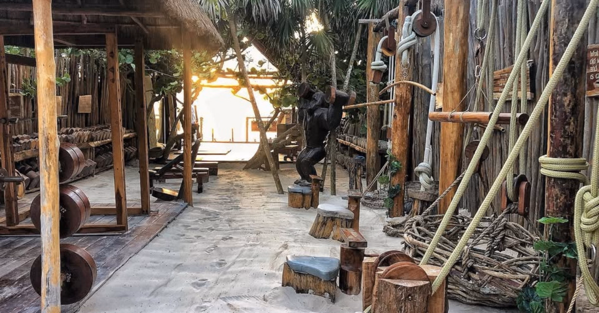 Outdoor gym in Tulum featuring wooden and rope equipment on sandy ground, surrounded by palm trees and rustic wooden structures, with sunlight streaming through the background.