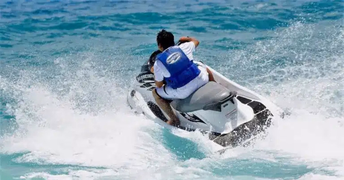 A person wearing a blue life jacket rides a white Jet Ski Tulum through choppy blue ocean waves, creating splashes of water behind them.
