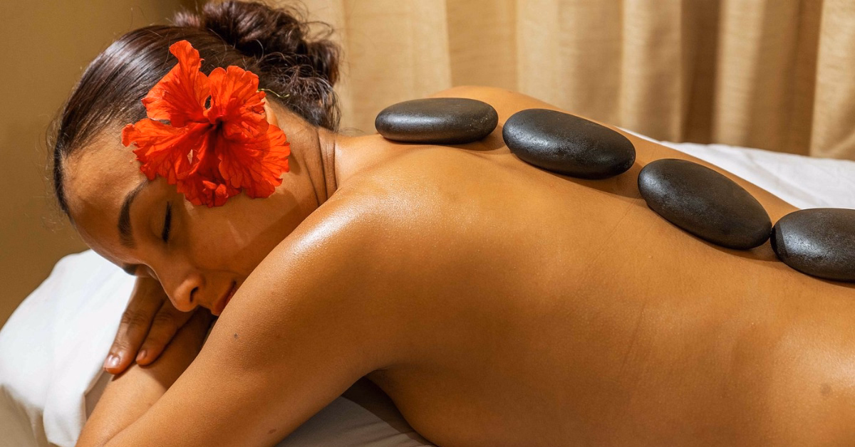A woman with a red flower in her hair lies face down on a massage table, eyes closed, with smooth black stones placed along her back, enjoying one of the relaxing hot stone massages in Tulum.