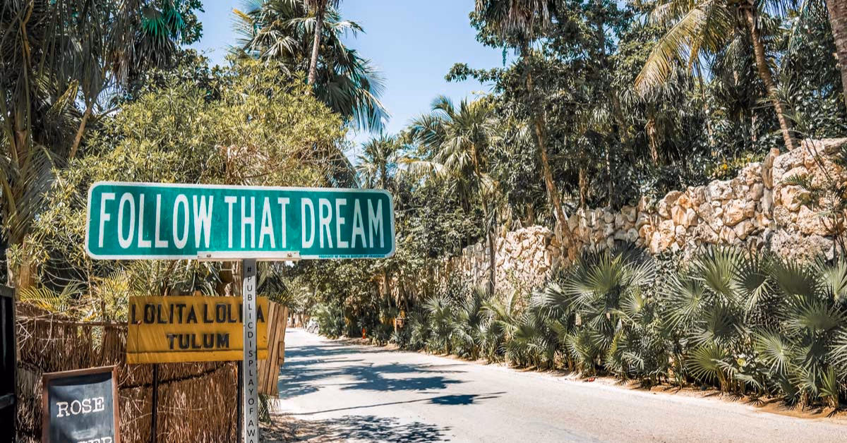 A green street sign saying FOLLOW THAT DREAM stands by a palm-lined road and stone wall beneath a clear blue sky in Tulum.