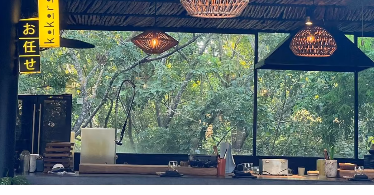 Open-air kitchen with wooden counters, woven lamps, glass wall to green trees, a yellow sign in Japanese, and inviting décor.
