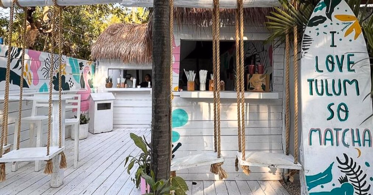 A small, white outdoor café with swings as seats, tropical plants, and colorful wall art. Known among healthy restaurants for its laid-back, beachy vibe, a surfboard sign reads, “I LOVE TULUM SO MATCHA.”.